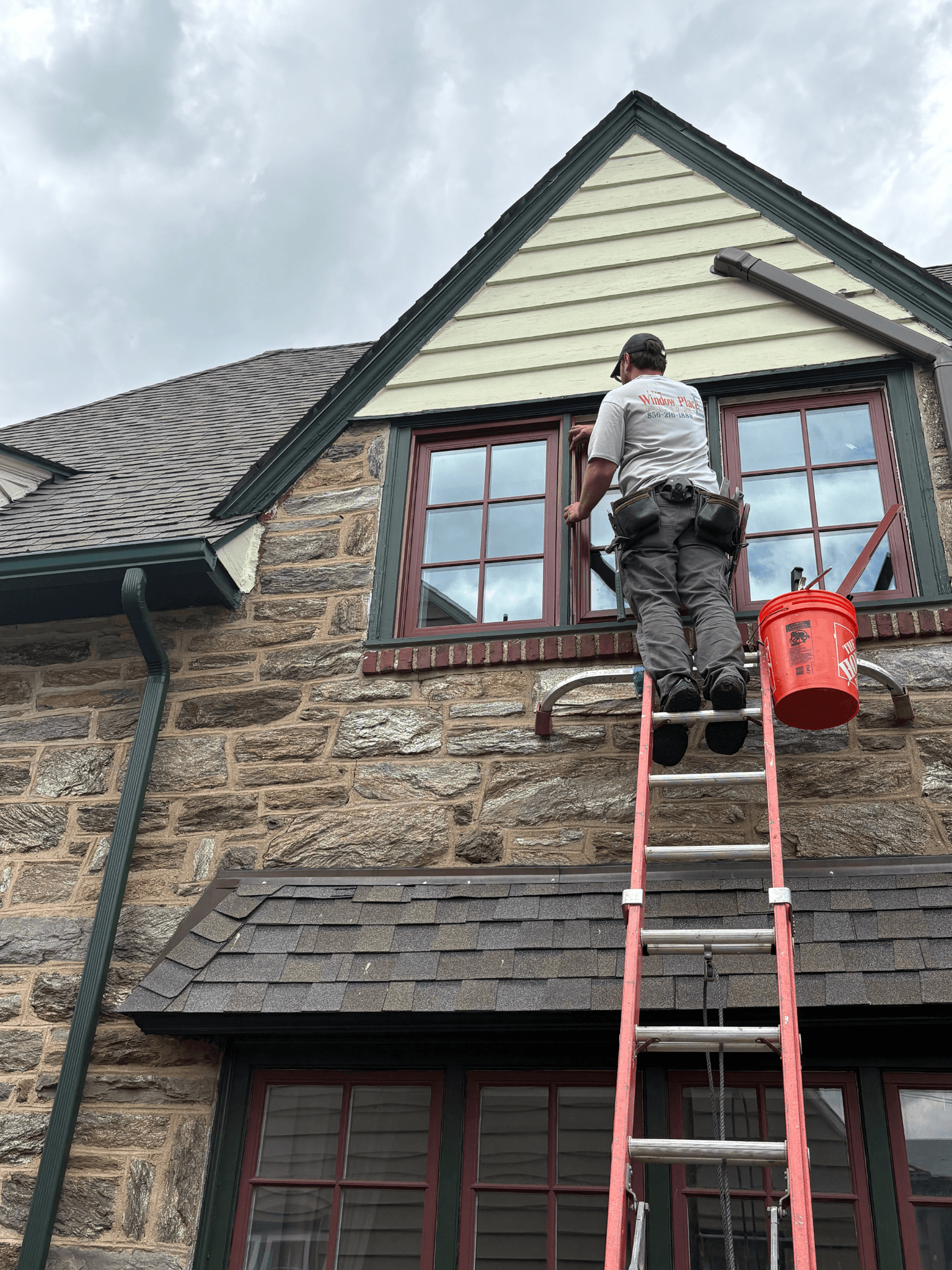 A worker on a ladder cleans second-story windows of a stone house. He wears a cap and uniform; an orange bucket hangs nearby. The sky is overcast.