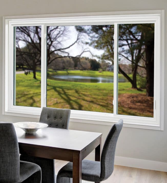 Dining area with gray upholstered chairs and a wooden table, set before a large window showcasing a serene landscape with green grass, trees, and a pond.