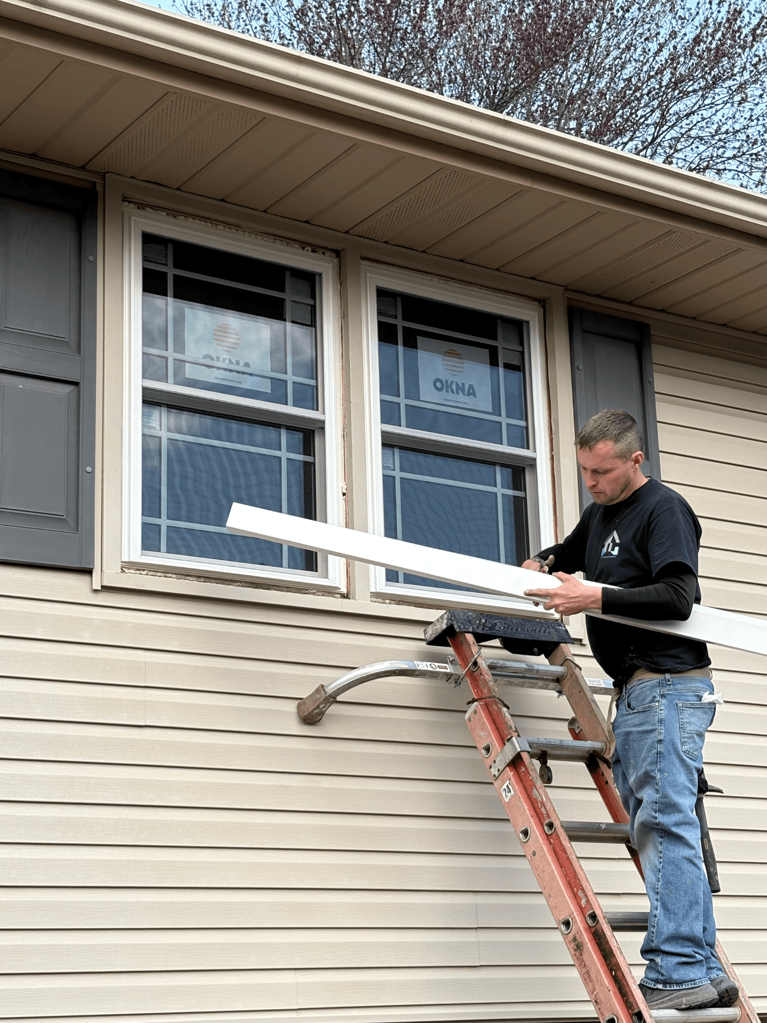 Man on ladder installing white trim under double windows on beige siding house; focused expression, clear day, branches in background.