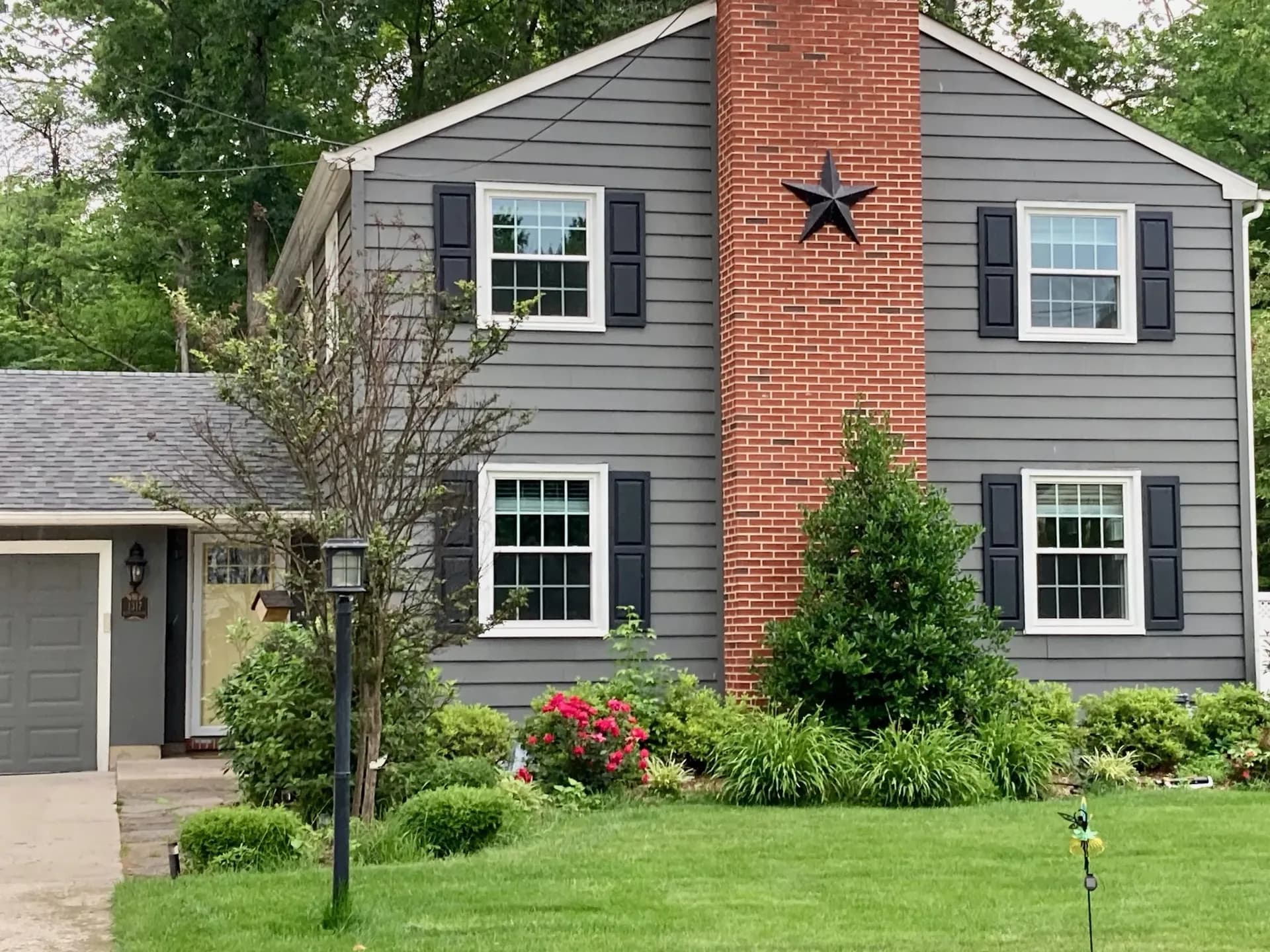 A gray two-story house with black shutters and a red brick chimney featuring a black star. Lush green lawn and vibrant bushes in front.