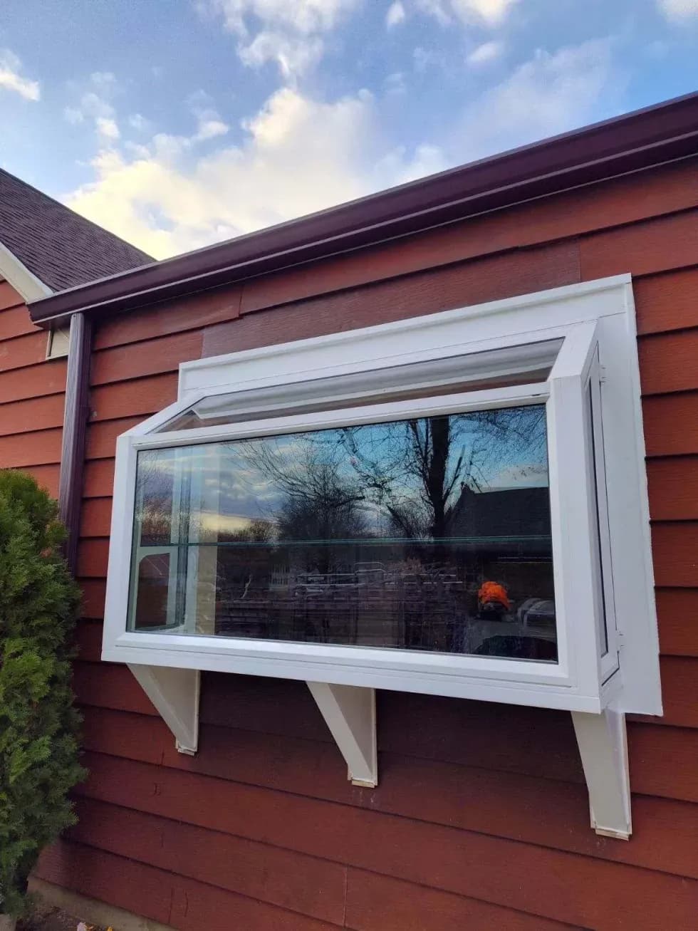 A rectangular glass bay window protrudes from a red wooden wall, reflecting the cloudy sky and bare trees. The mood is calm and serene.