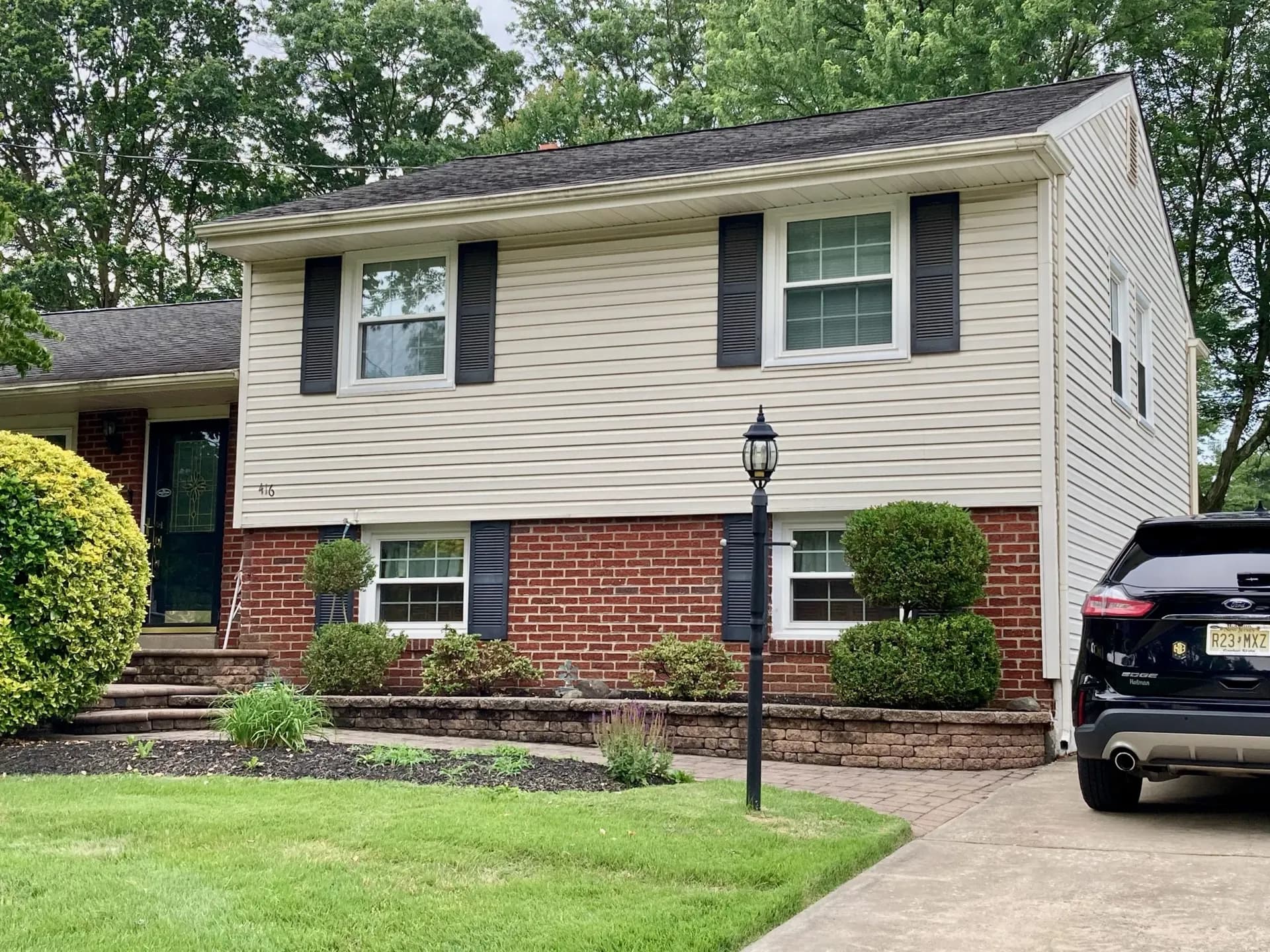 Suburban split-level house with brick and siding exterior, black shutters, and a well-kept lawn. A driveway with a parked car is on the right.
