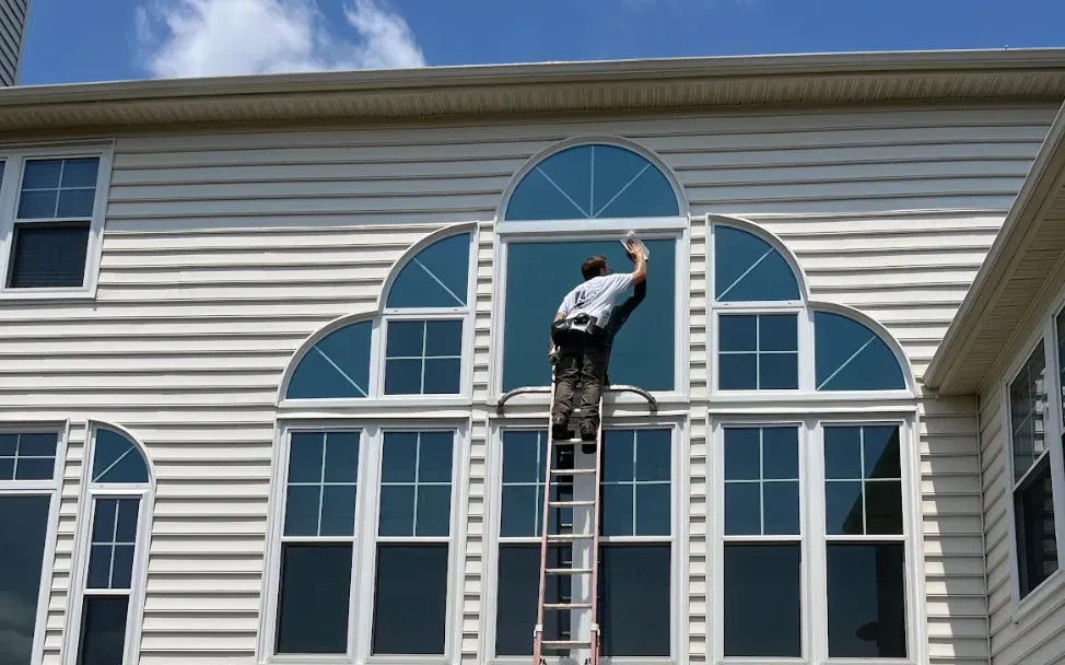 A person on a ladder cleans a large window with an arched design on a two-story house exterior, under a clear blue sky with a few clouds.