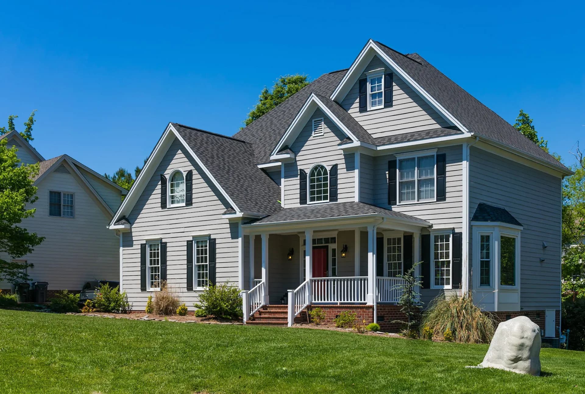 Two-story gray house with white trim and a red door, featuring large windows and a front porch with white railing. Surrounded by a green lawn and trees under a clear blue sky.