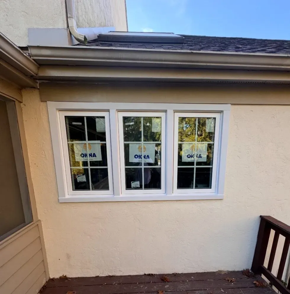 Newly installed triple window with white trim on a beige stucco wall, reflecting a clear blue sky. Wooden deck below, conveying a fresh, clean look.