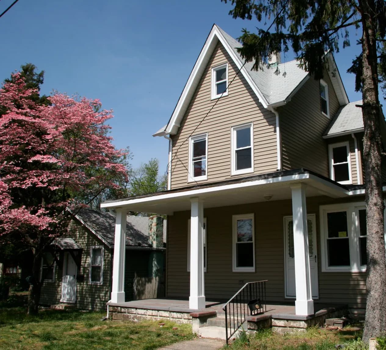 A brown, two-story house with white trim and a porch stands under a clear blue sky. A blossoming pink tree adds a touch of spring to the scene.