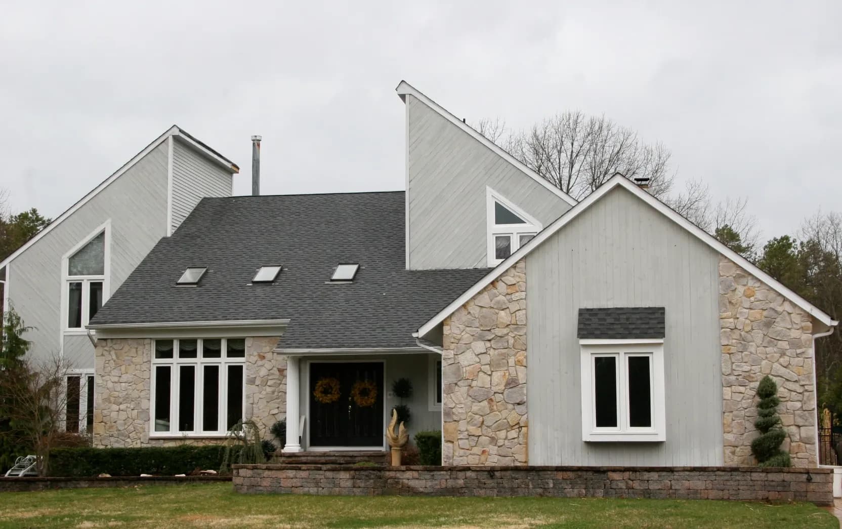 Modern two-story house with a unique angular roof design, stone and beige siding, large windows, and a small front garden under an overcast sky.