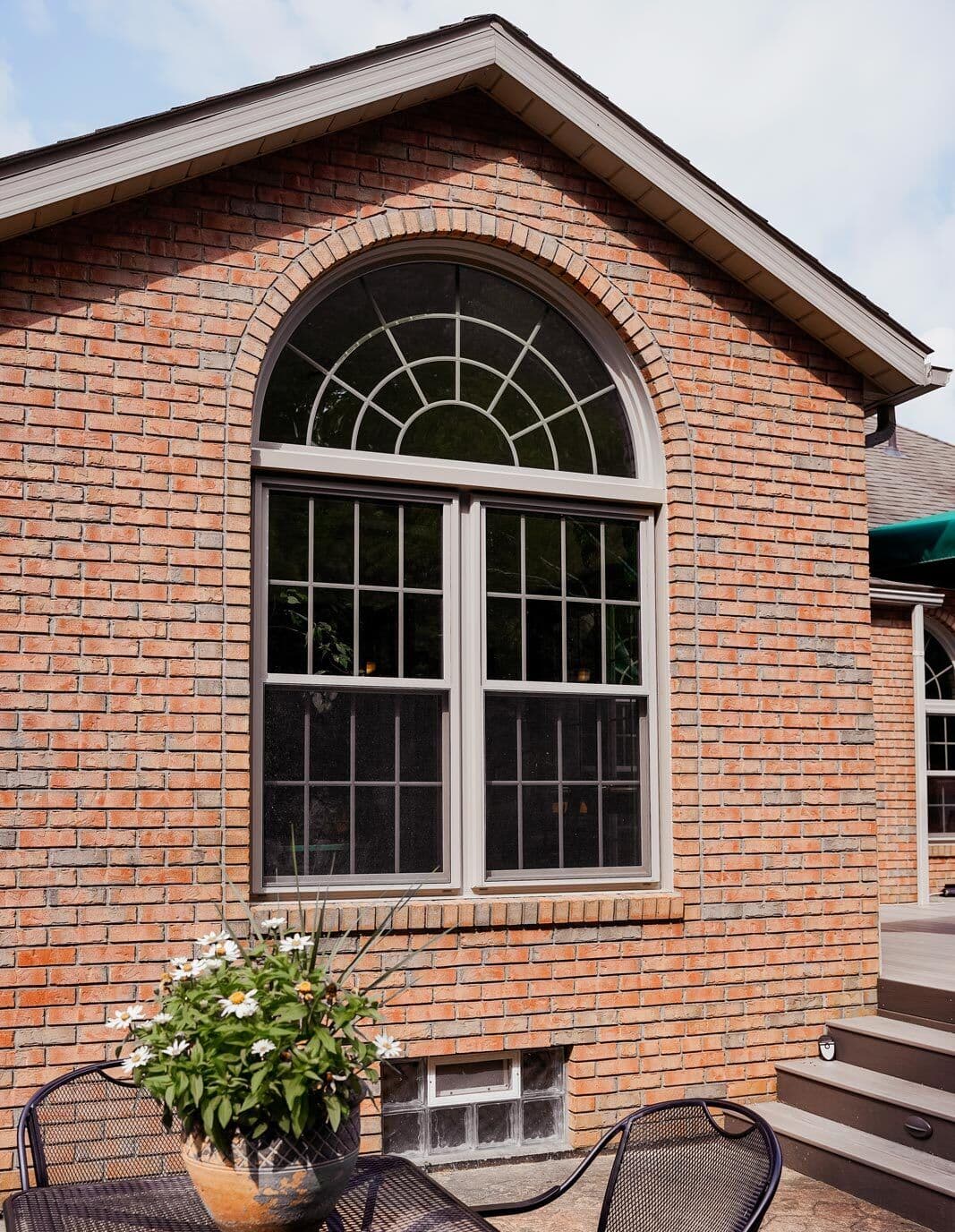 Brick house exterior with a large arched window, reflecting sunlight. Below is a patio table with a potted plant, exuding a peaceful, sunny atmosphere.