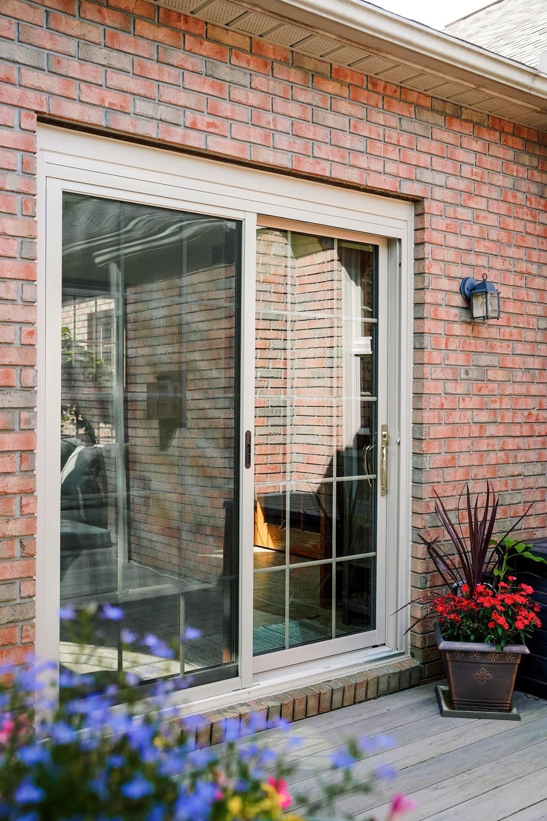 Sliding glass door on a brick wall leads to a cozy interior. Colorful flowers and a decorative plant on a wooden deck add a vibrant, inviting touch.