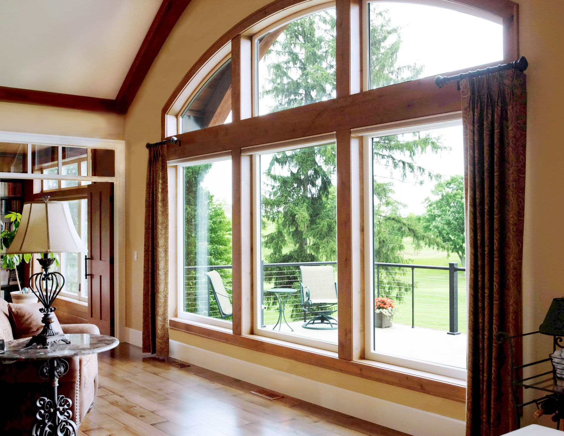 Spacious living room with large arched windows overlooking a lush green landscape. Elegant curtains frame the view, creating a serene, airy ambiance.
