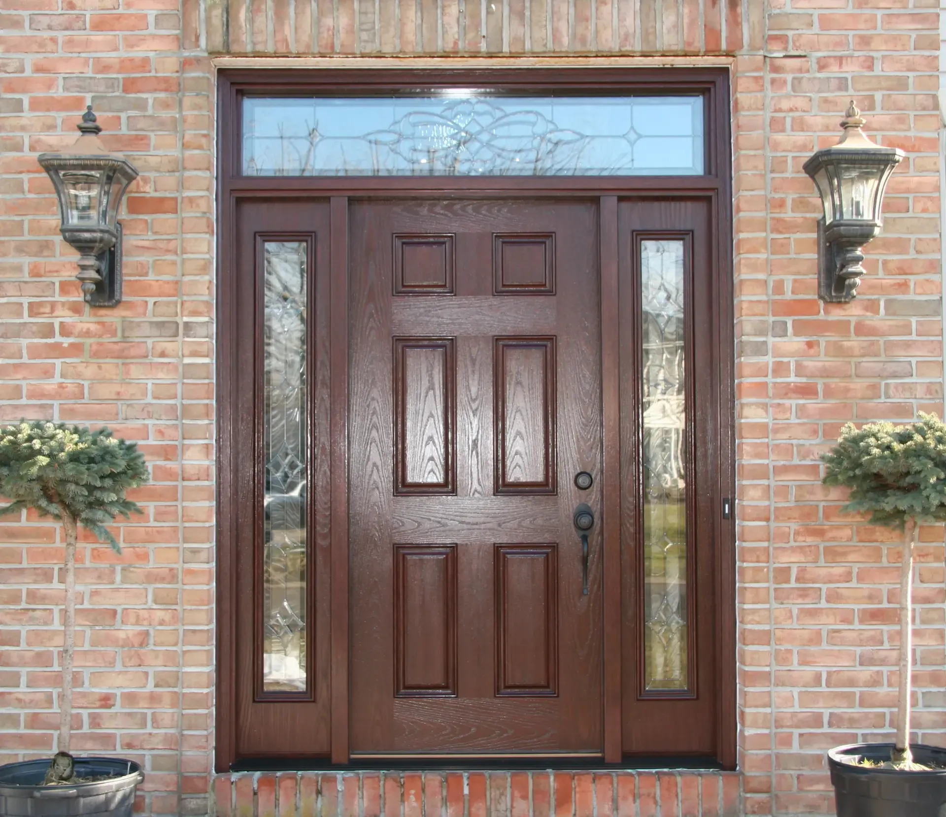 A dark brown wooden front door with decorative glass panels is flanked by two sconces and potted shrubs, set against a red brick wall.