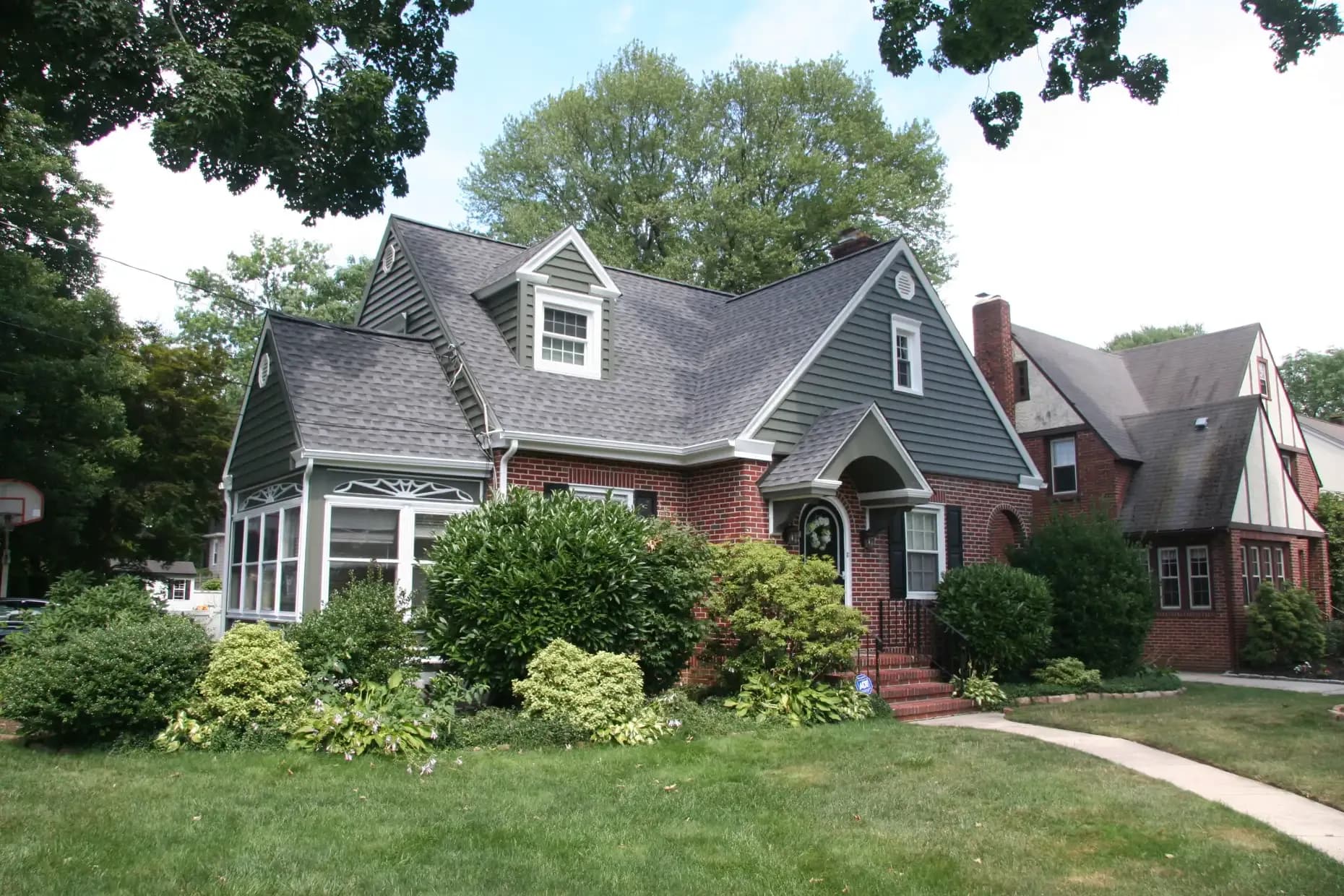 Quaint brick house with a gabled roof and dormer window, surrounded by lush green bushes and a neatly maintained lawn, conveying a cozy, welcoming feel.