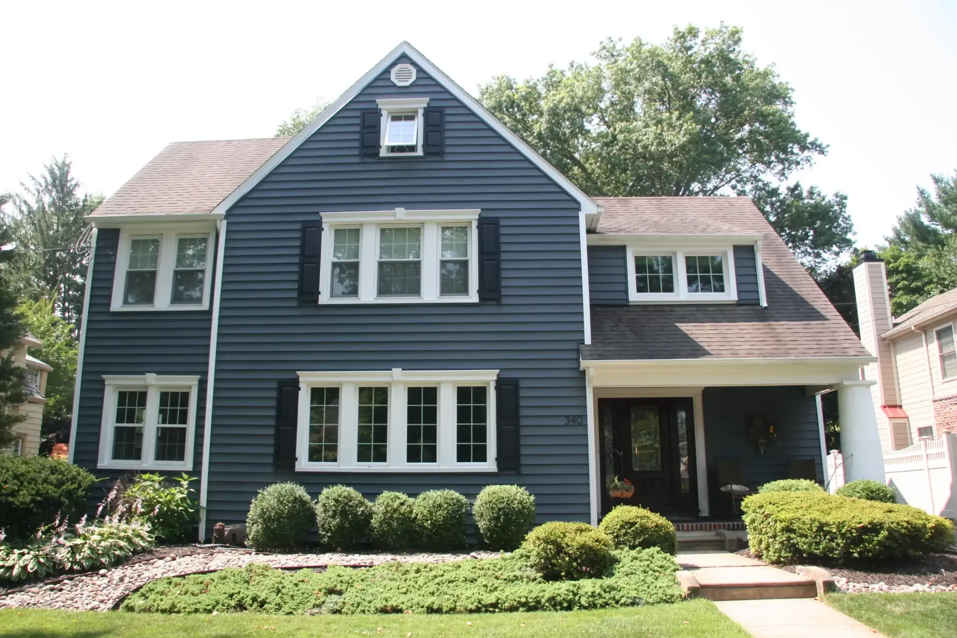 A charming two-story blue house with white trim, large windows, and black shutters. It's framed by manicured shrubs and a lush green lawn under a clear sky.