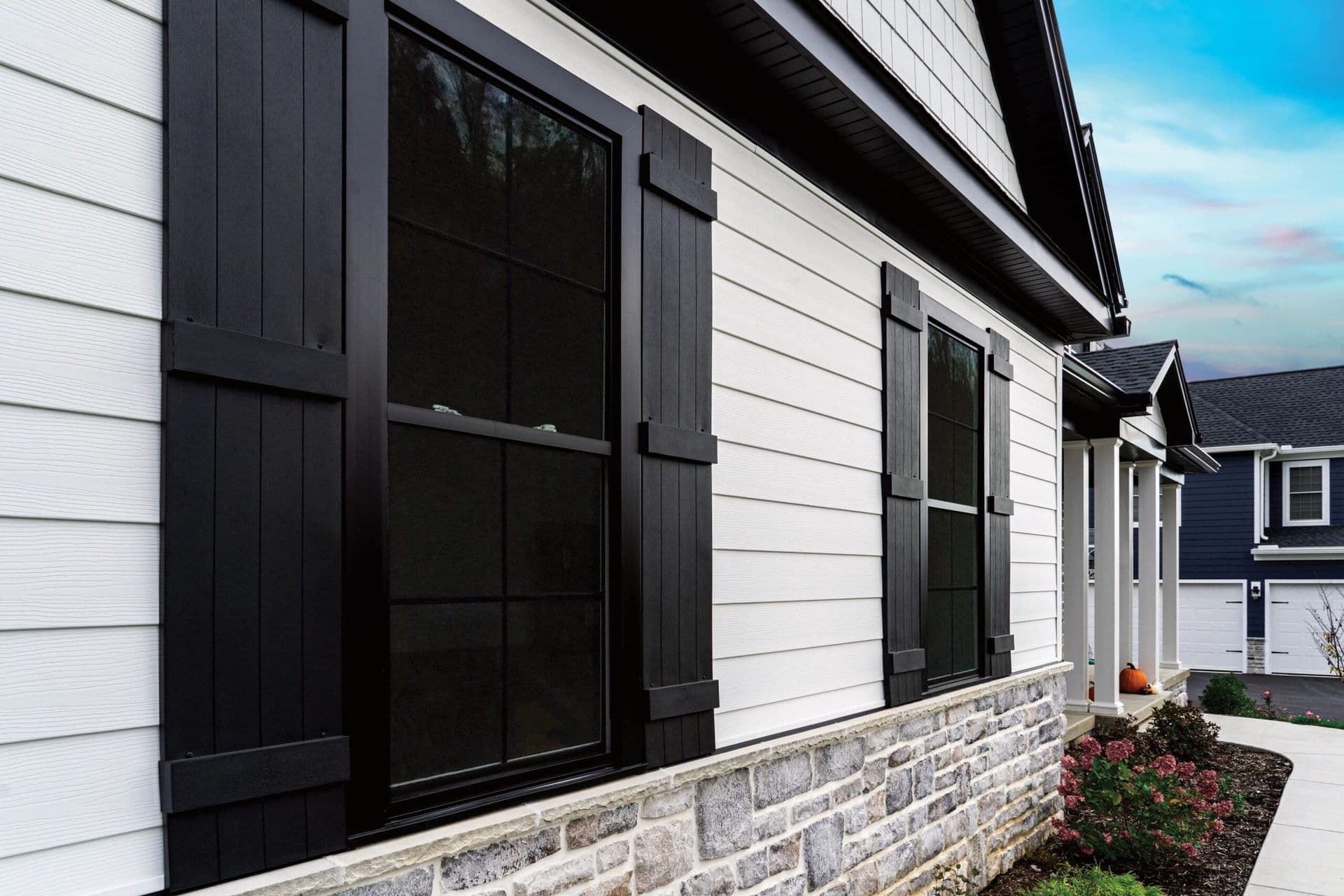 Close-up of a modern house exterior with black window shutters, white siding, and stone accents. A small garden and clear sky enhance the calm ambience.