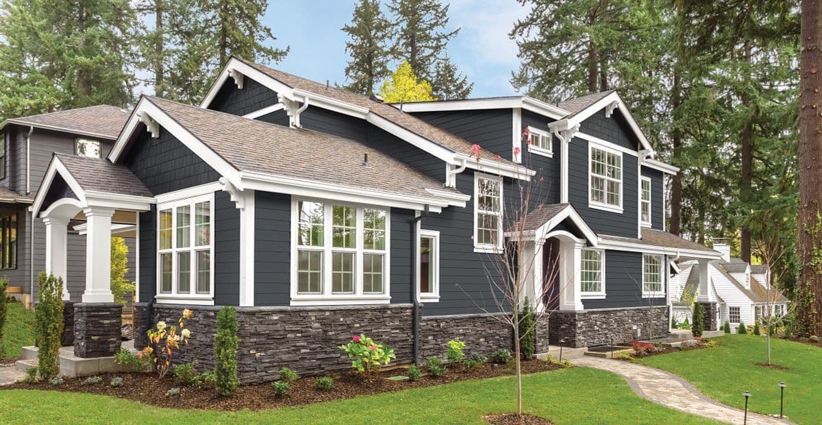 A modern, two-story house with dark blue siding and white trim, surrounded by lush greenery and tall trees, under a clear sky, exuding elegance.