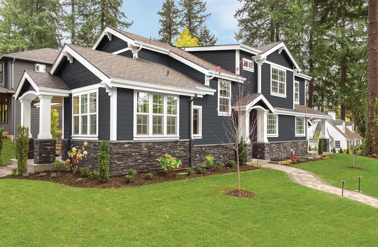 A modern, two-story house with navy blue siding and stone accents sits amid lush green grass. Tall trees surround the property under a clear sky.