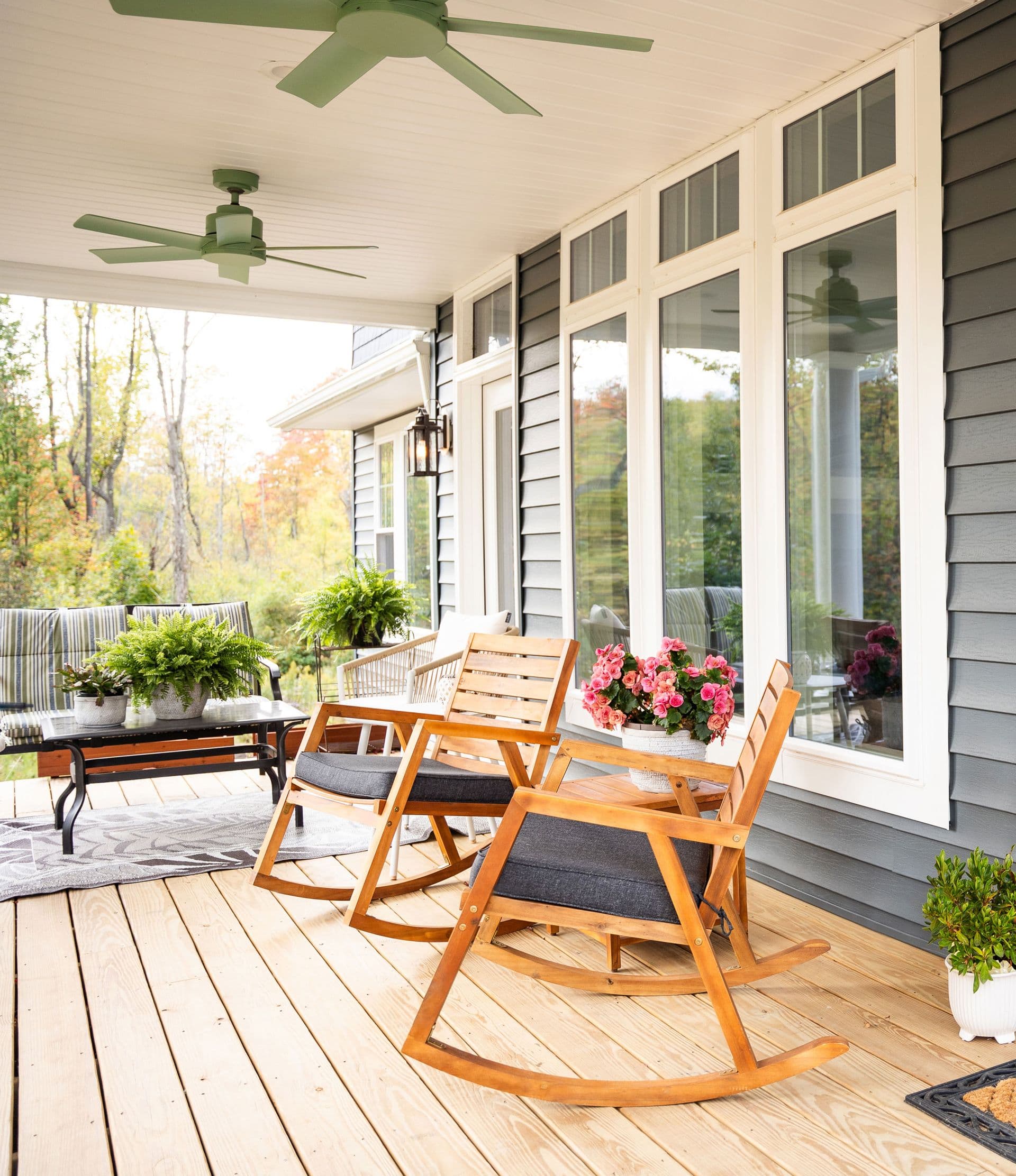 Cozy porch with wooden rocking chairs, vibrant pink flowers, and potted plants. Gray siding, large windows, and overhead fans add a serene, inviting feel.