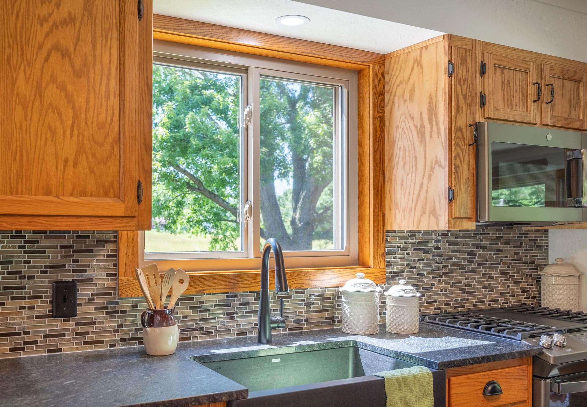 Kitchen scene featuring wooden cabinets, a window with tree view, black countertop, green sink, utensils in a jar, and a stainless steel stove.