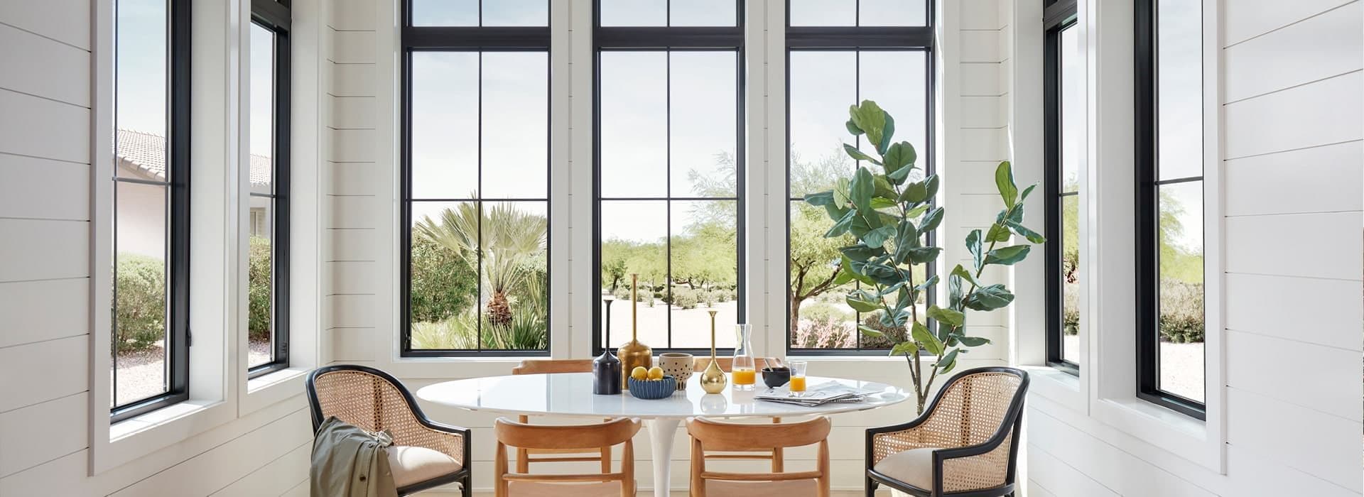 Bright dining area with large black-framed windows, a round table with four wooden chairs, a potted plant, and dishes with fruit. Airy and inviting.