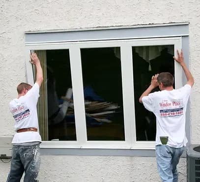 Two workers are installing a large white-framed window on a beige wall. Both wear matching branded shirts and appear focused and cooperative.