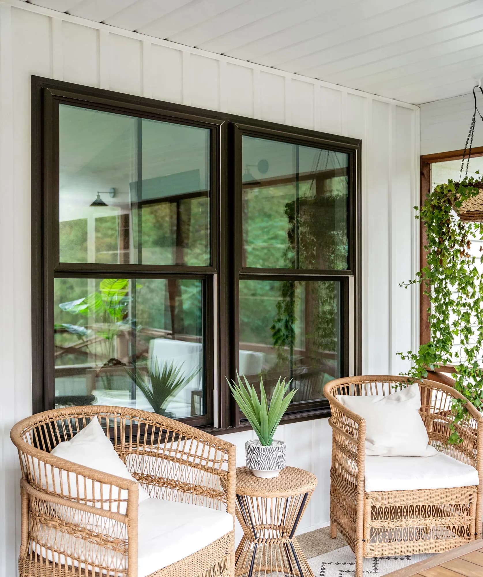 Cozy patio with two wicker chairs, white cushions, and a small table with a plant. Large windows and lush greenery enhance the serene atmosphere.