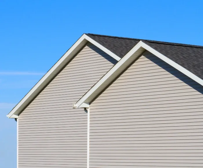 Side view of two adjacent beige house rooftops against a clear blue sky. The buildings have matching gable roofs and vinyl siding, conveying suburban calm.