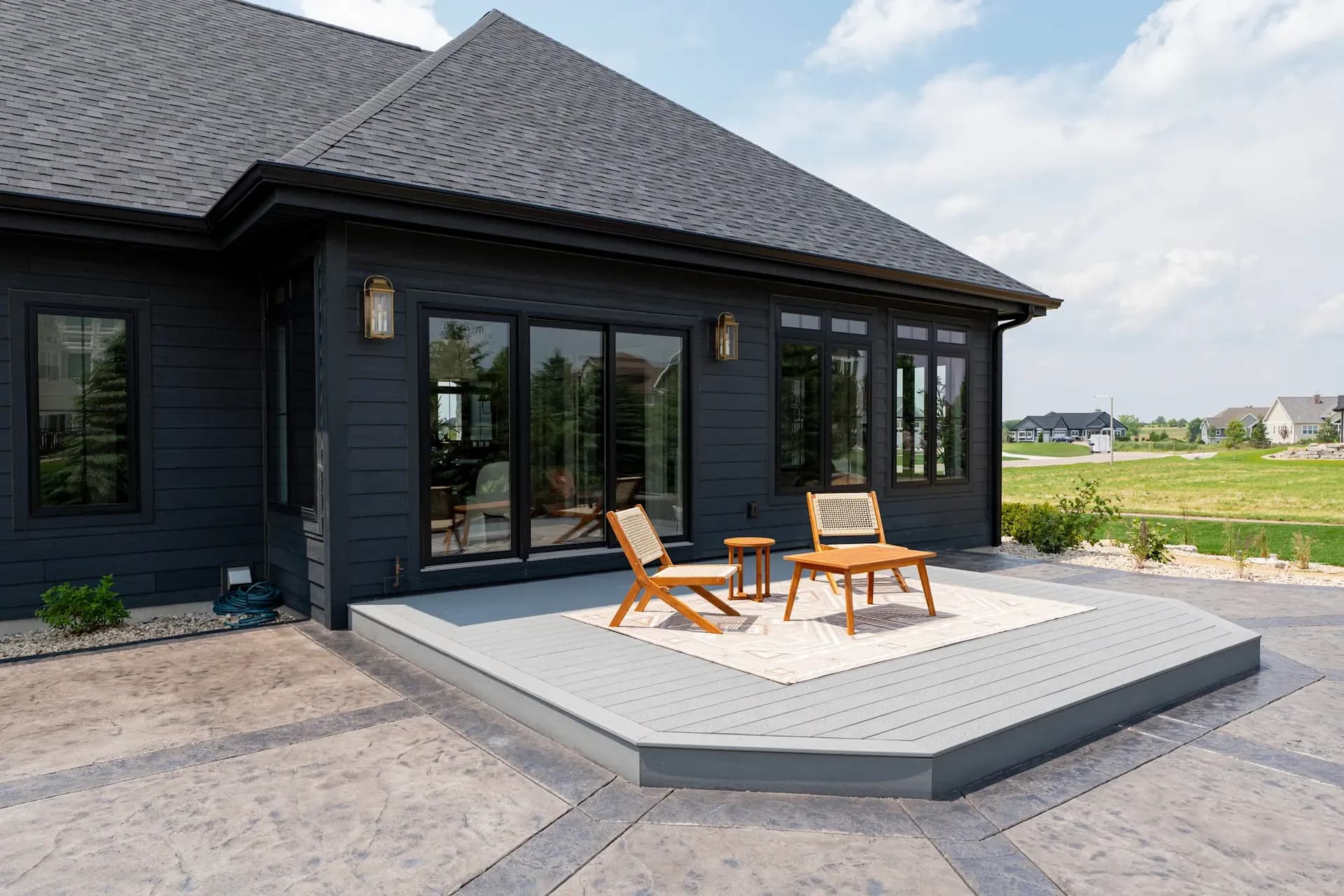 Modern patio with two wooden chairs and a small table on a gray deck. A dark house with large windows is in the background, set against a sunny sky.