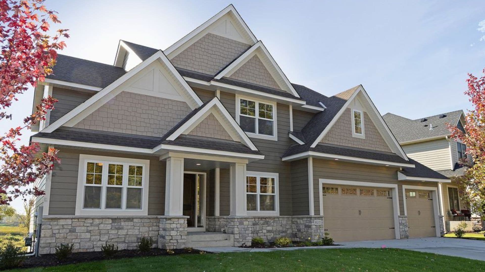 A two-story house with taupe siding, stone accents, and a gabled roof. The front features large windows, a manicured lawn, and surrounding red trees.