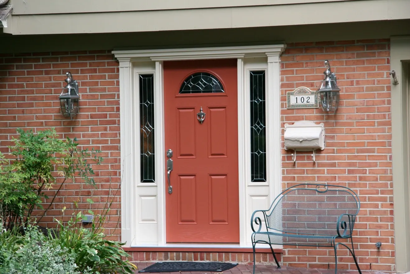 A red front door with white trim and decorative glass panels is centered on a red brick wall. Two lanterns flank the door, with a metal bench and plants nearby.