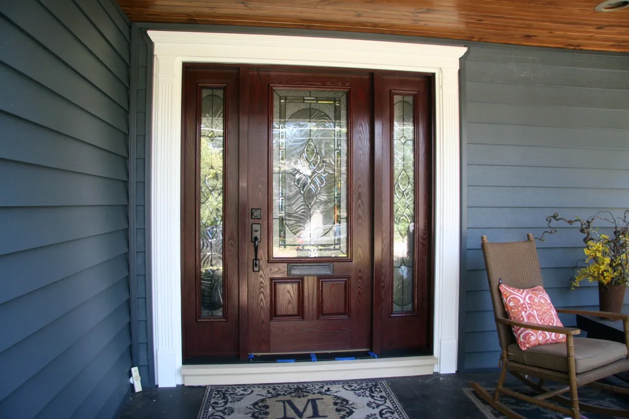 A dark wooden front door with decorative glass panels is flanked by blue siding. A rocking chair with a decorative pillow and flowers sits nearby, creating a welcoming porch scene.