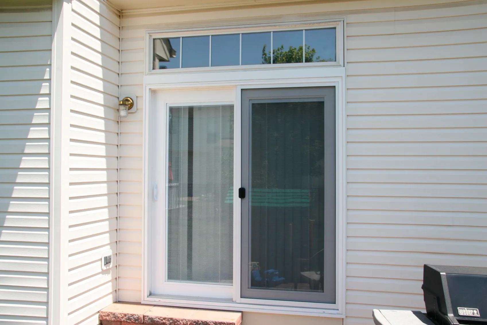 Sliding glass patio door with blinds, set in a white siding wall. Sunlit, with a small transom window above and a metal outdoor light fixture.