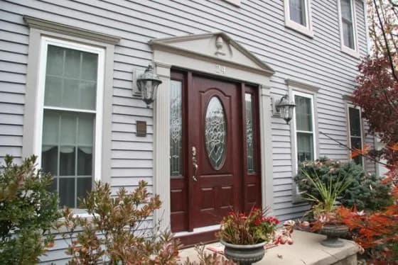 Elegant home entrance with dark red door featuring intricate oval glass design. Flanked by tall windows and lanterns, surrounded by autumnal plants.