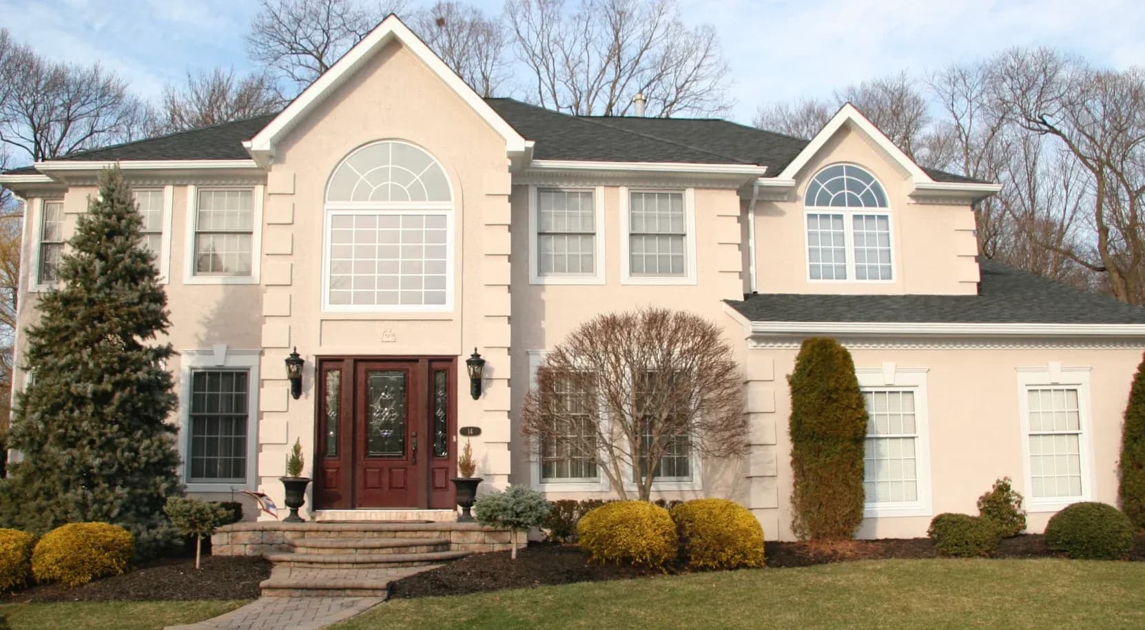 Two-story beige house with a dark roof, large arched windows, and a red front door. Neatly trimmed shrubs and trees decorate the front yard.
