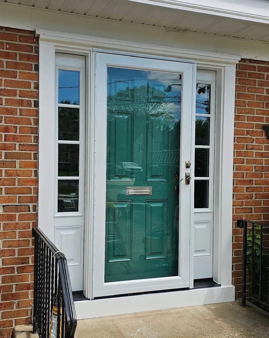 A teal front door with a rectangular glass pane stands between red brick walls, framed by white trim and side windows, reflecting a tree and sky.