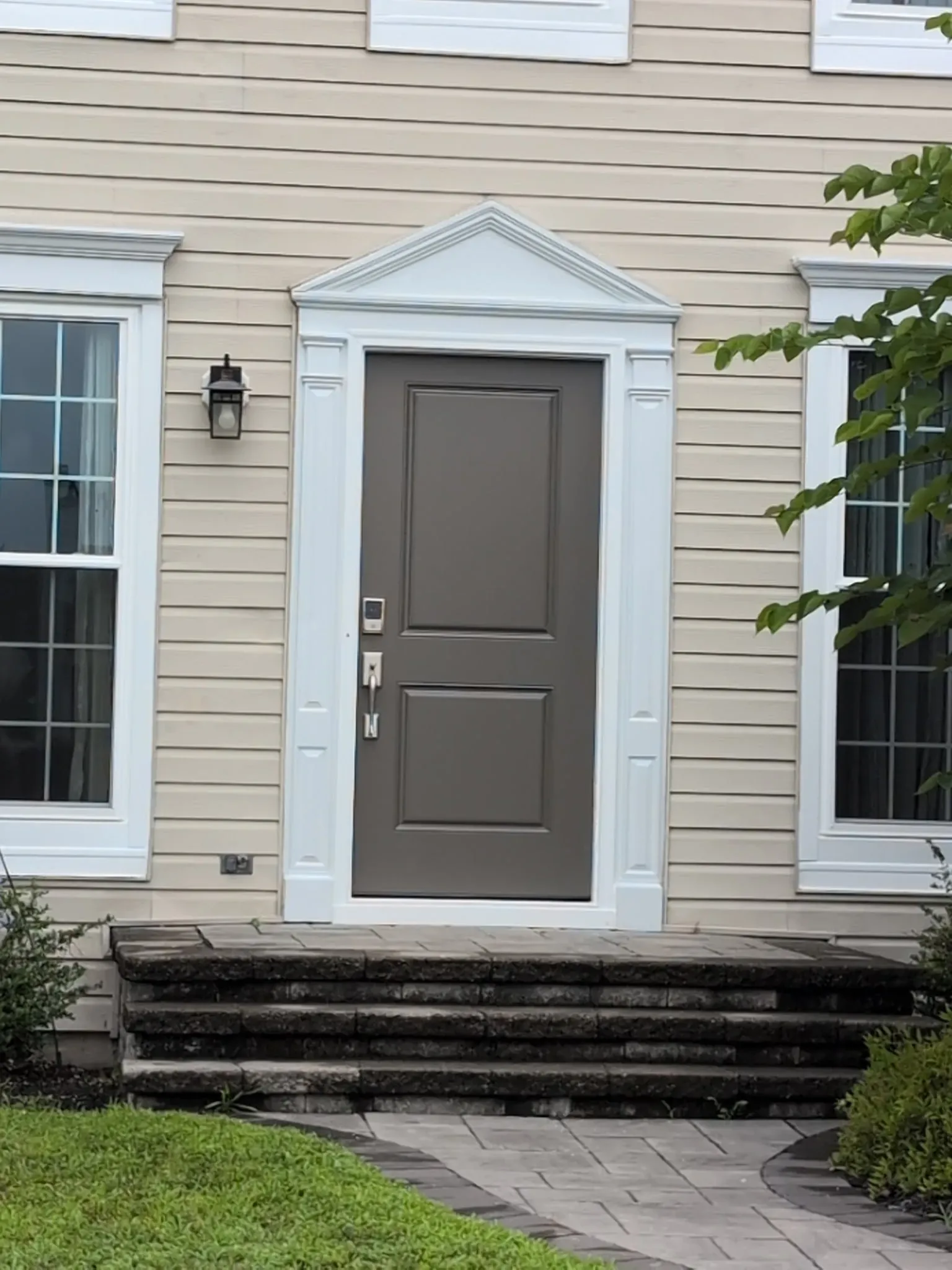 A beige house features a gray front door with white trim, flanked by two large windows. A black lantern is on the left, and stone steps lead to the entry.