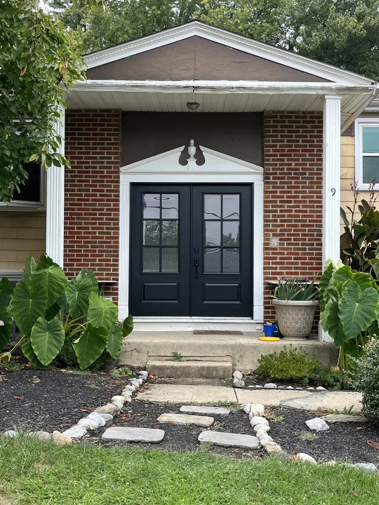 Front of a red brick house with black double doors and white framing, flanked by columns. Lush green plants and a stone path create a welcoming entrance.