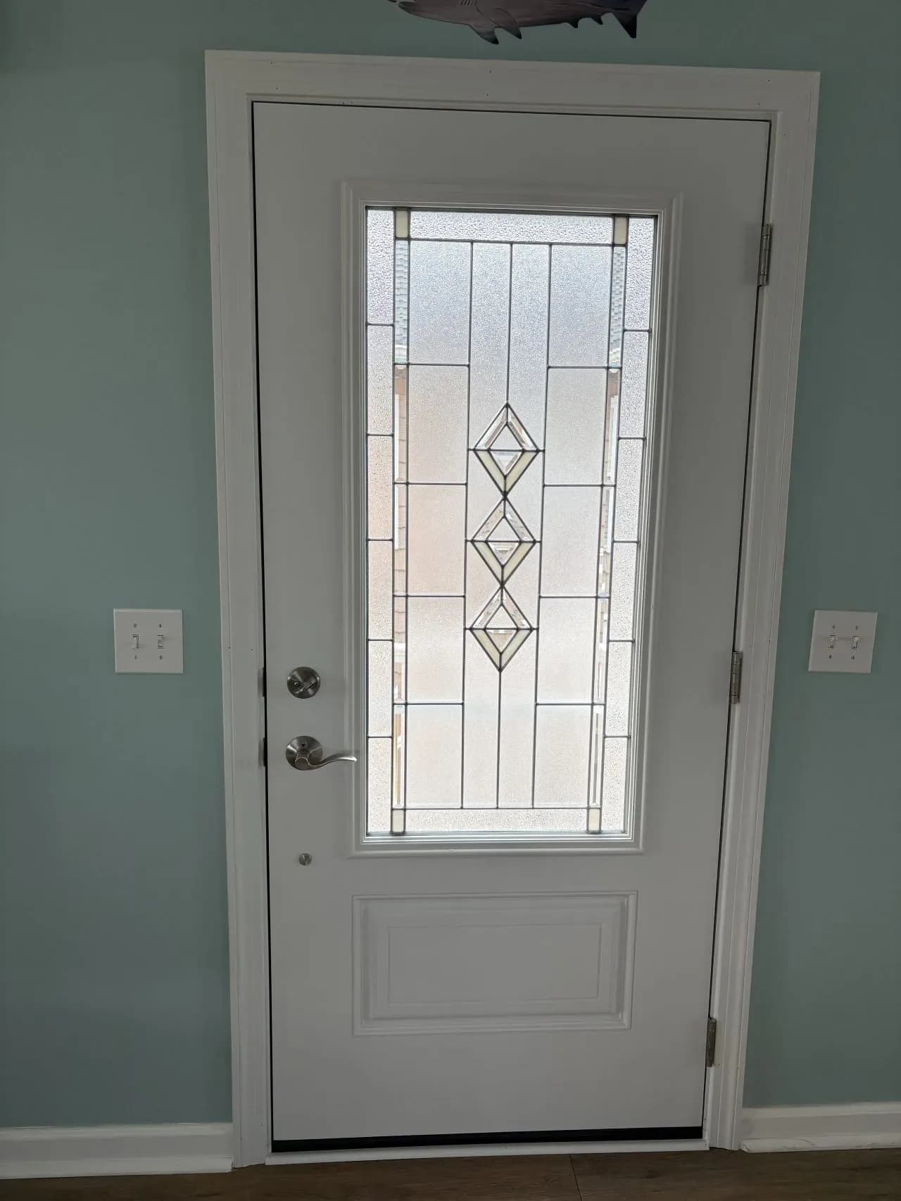White door with decorative glass panel in geometric pattern, set in a light blue wall. Two light switches are visible on each side. The atmosphere is calm.