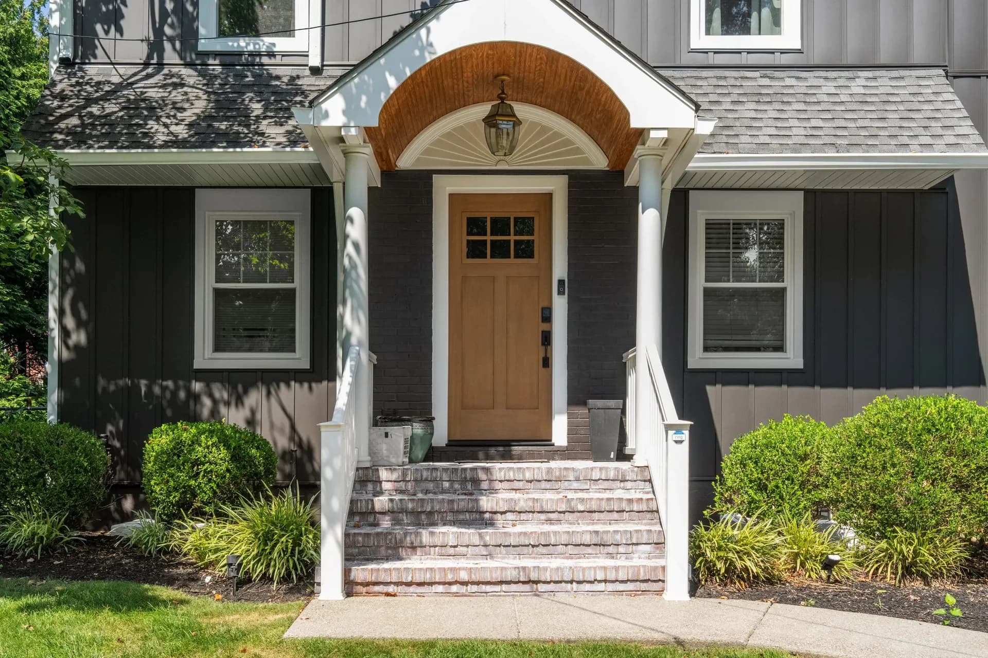 Front porch of a house with a wooden door, white columns, and brick steps. Surrounded by greenery, creating a welcoming and serene entrance.