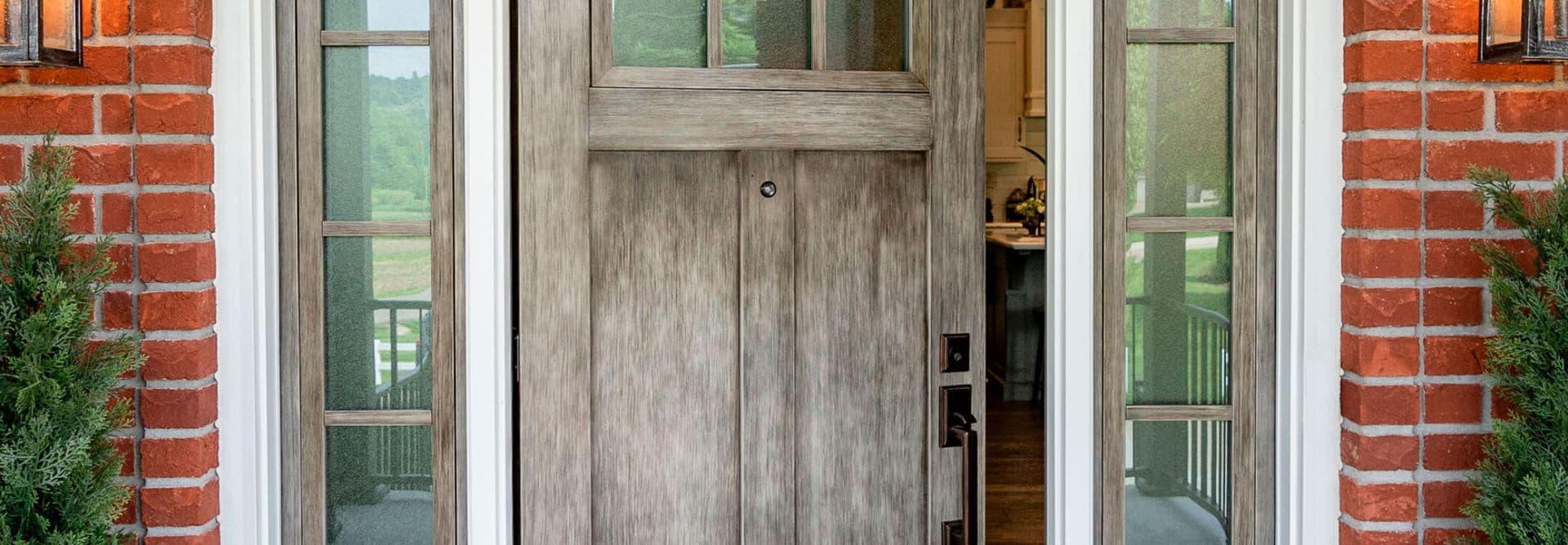 A rustic wooden front door, slightly ajar, set in a red brick wall with glass side panels. Greenery flanks the entrance, suggesting a welcoming home.