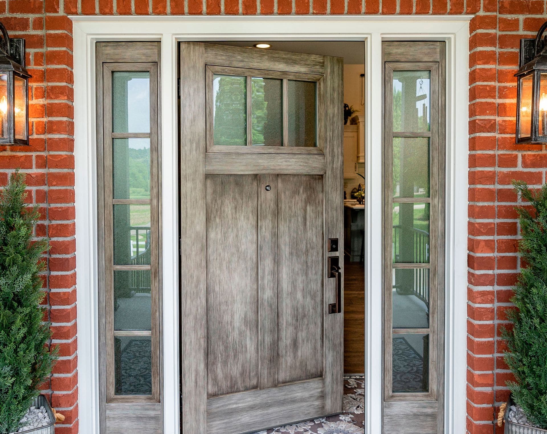 Rustic wooden front door ajar, flanked by tall side windows. Red brick exterior and warm lantern lights create a welcoming, cozy ambiance.