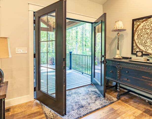 Open French doors lead to a sunlit deck, surrounded by lush greenery. The cozy room has a vintage blue sideboard and patterned rug, creating a serene vibe.