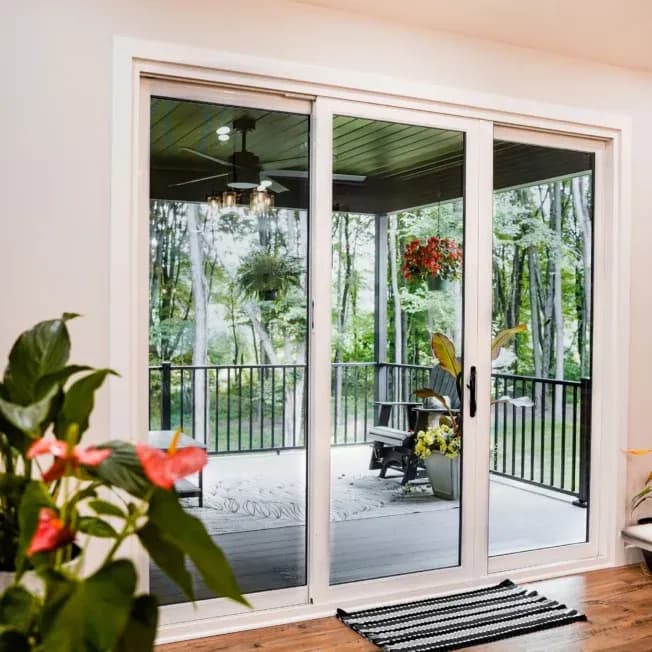Stylish glass doors open to a cozy porch with potted plants and chairs, overlooking lush green trees. A vibrant, inviting indoor-outdoor space.