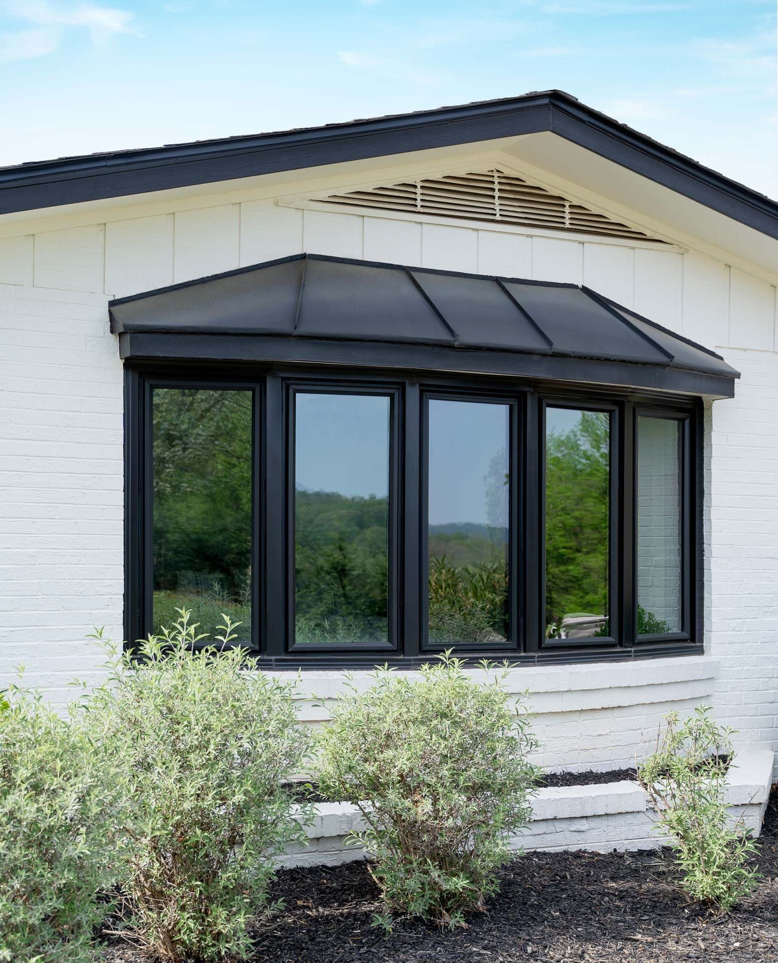 Modern black-framed windows on a white brick house with a sloped roof. Bushes in the foreground, blue sky in the background. Calm and inviting.