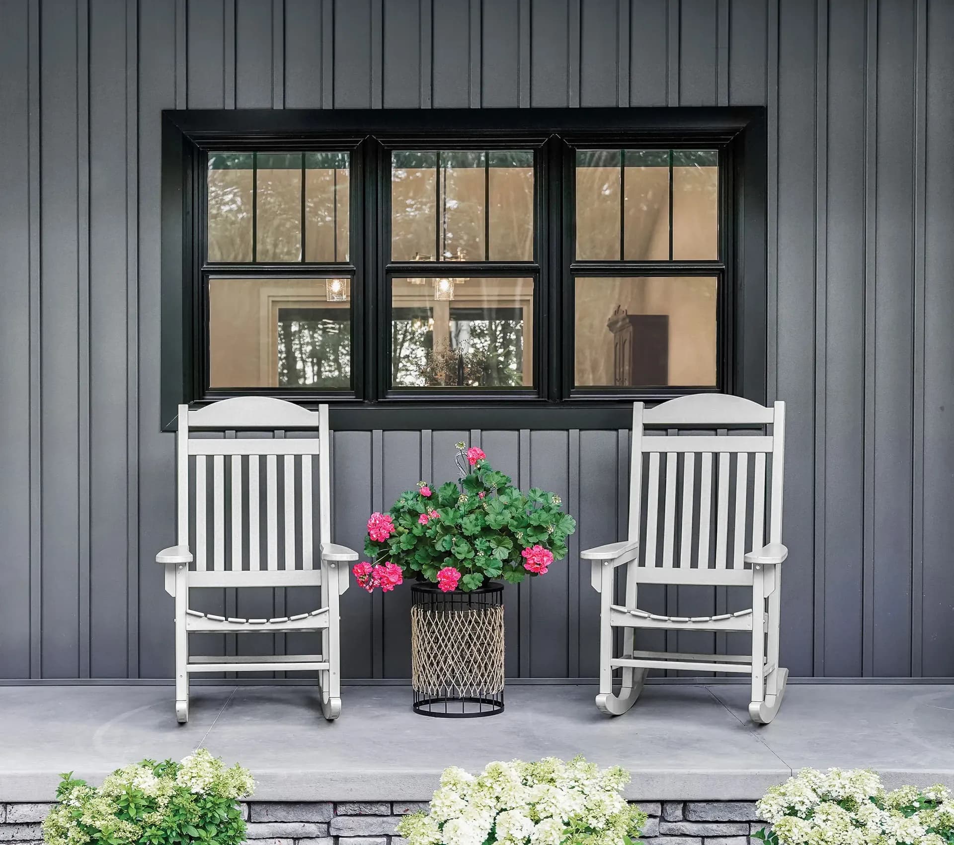 Two white rocking chairs sit on a patio, flanking a pot of blooming pink flowers. Behind them is a dark-paneled wall with a large window.
