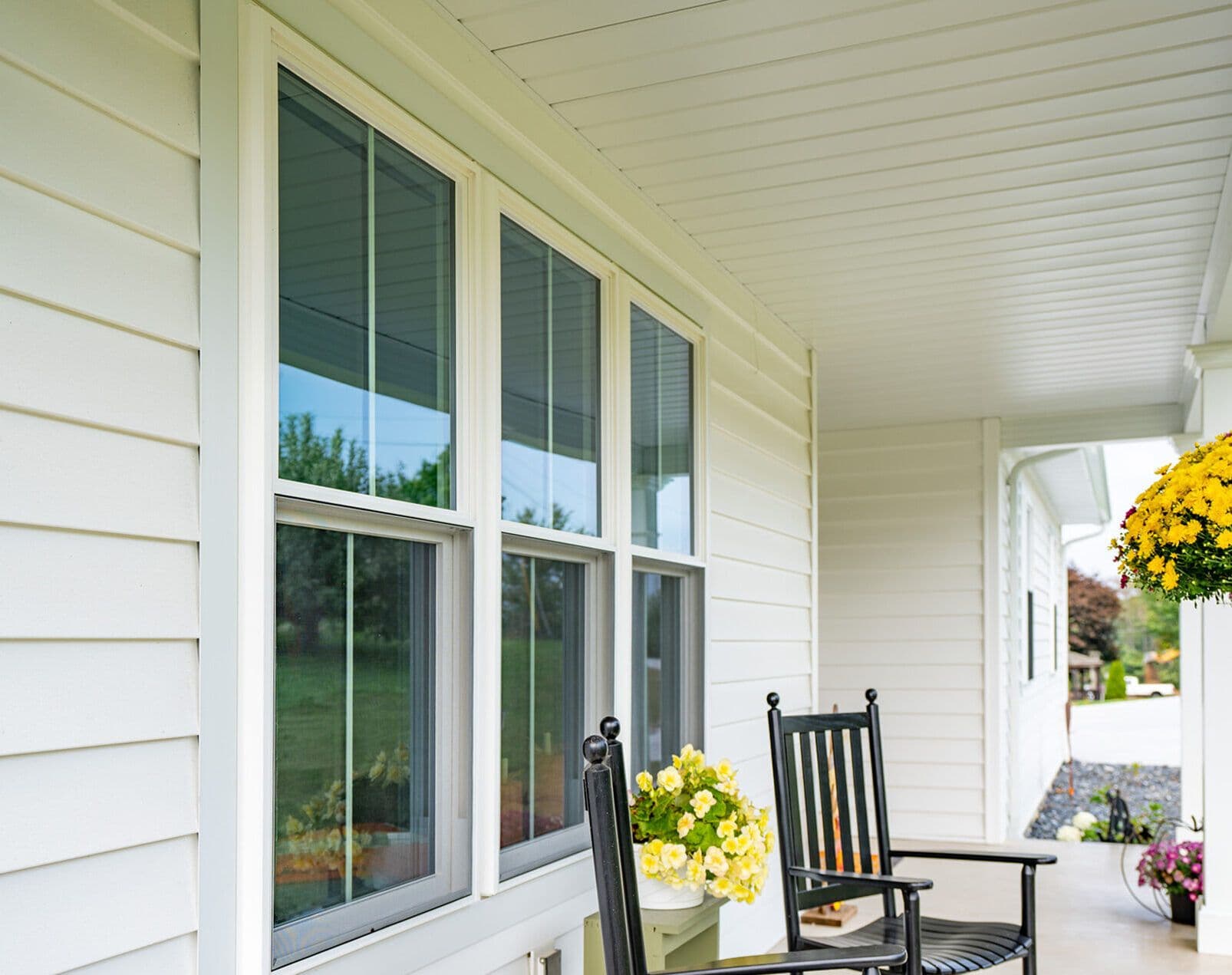 Front porch with two black rocking chairs next to large windows on a white house. Hanging basket with yellow flowers; sunny and inviting atmosphere.