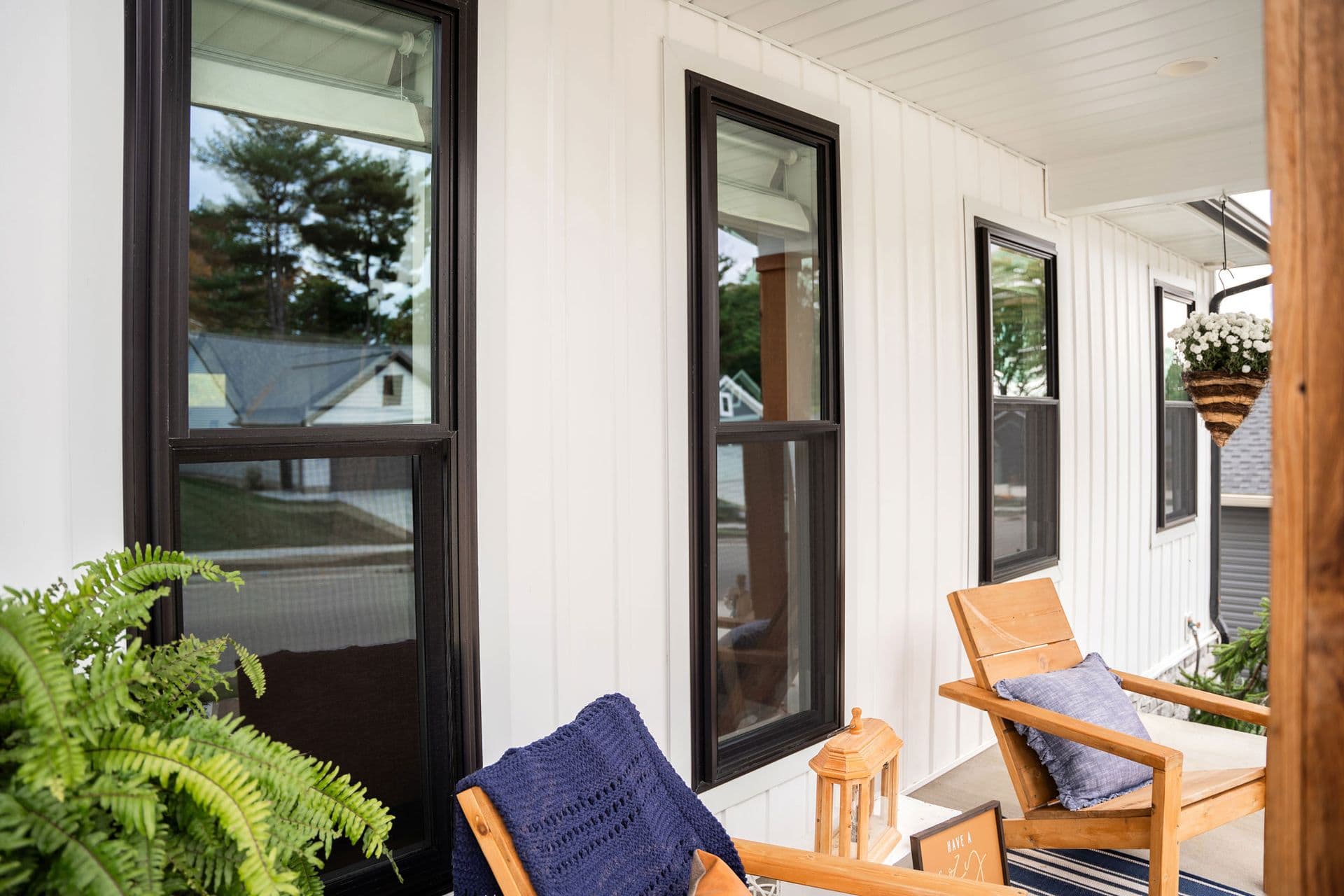 A cozy white porch with black-trimmed windows, wooden chairs, and blue cushions. Green plants and a hanging basket add a fresh, inviting feel.