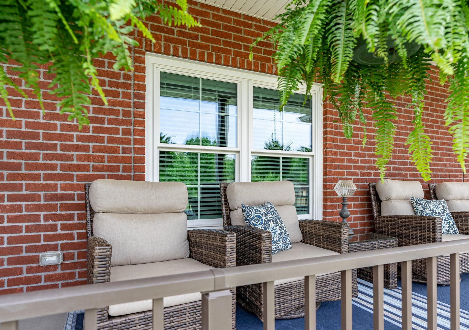 Front porch with wicker chairs, beige cushions, and patterned pillows. Hanging ferns add greenery. Brick wall backdrop creates a cozy, inviting feel.