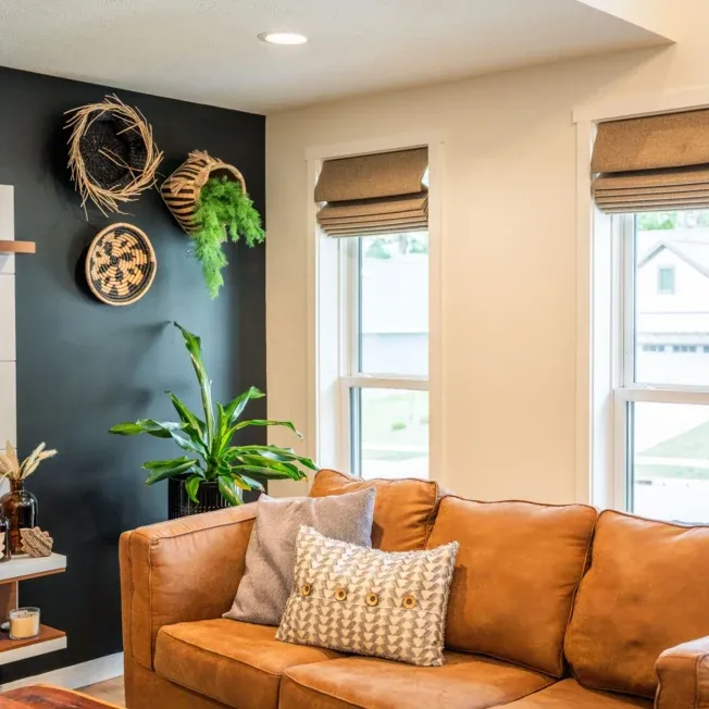 Cozy living room with a tan sofa, patterned cushions, and indoor plants. A black accent wall showcases woven baskets and provides a warm, inviting atmosphere.