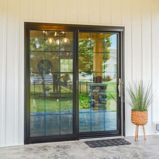 Sliding glass door with a black frame reflecting an outdoor scene of trees and a house. A potted plant sits nearby on a patterned floor.