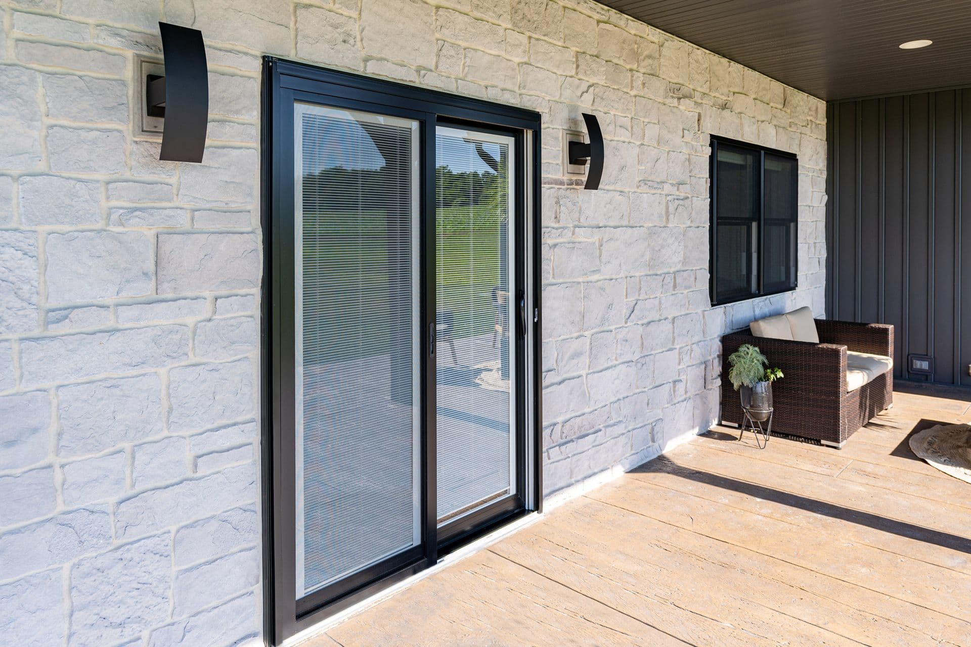 Covered patio with stone walls features sliding glass doors and wicker seating. Warm, inviting atmosphere with greenery and modern wall lights.